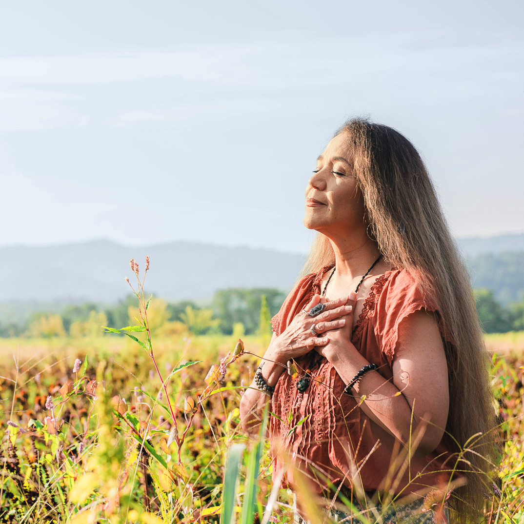Woman with long hair enjoying nature, hands on heart, in a field with mountains and blue sky in the background.