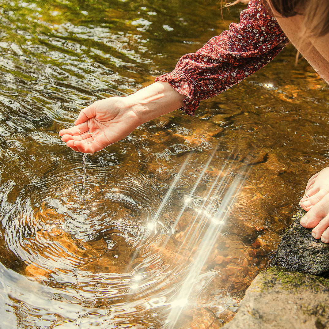 Person gently touching the surface of a calm stream, creating ripples in the sparkling water.