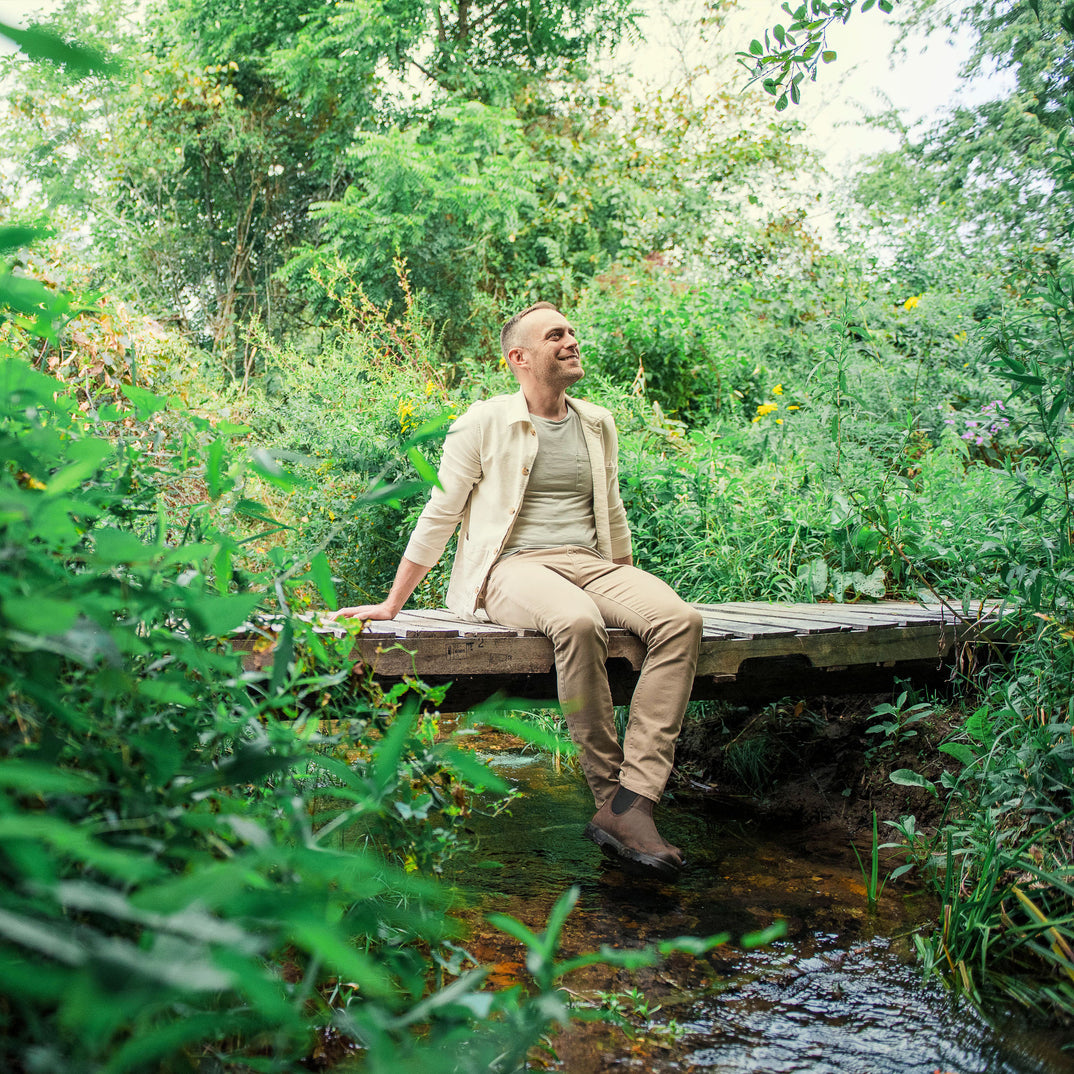 Man sitting on a wooden bridge over a stream, surrounded by lush greenery and summer flowers.